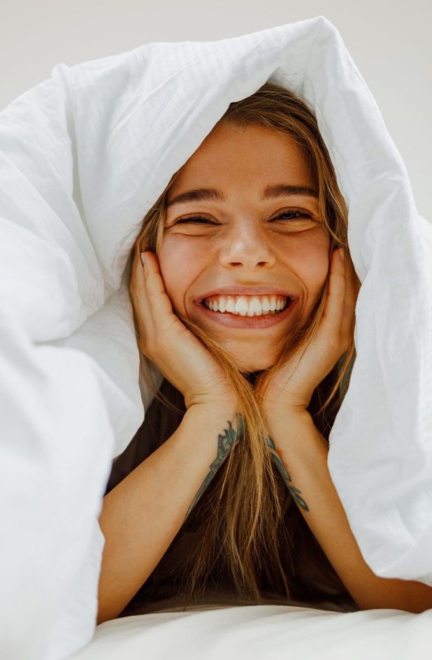 A,Woman,Lays,In,Bed,Under,A,White,Blanket,,Smiling